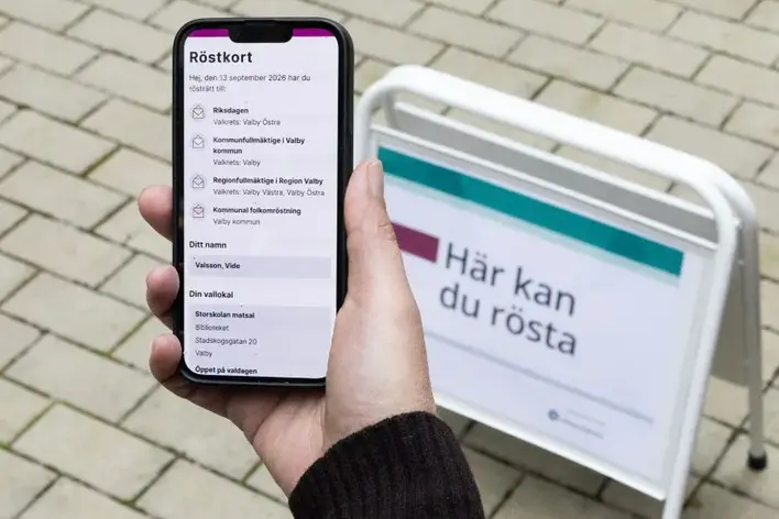 Hand som håller i mobiltelefon med digitalt röstkort och en skylt som visar: här kan du rösta.
